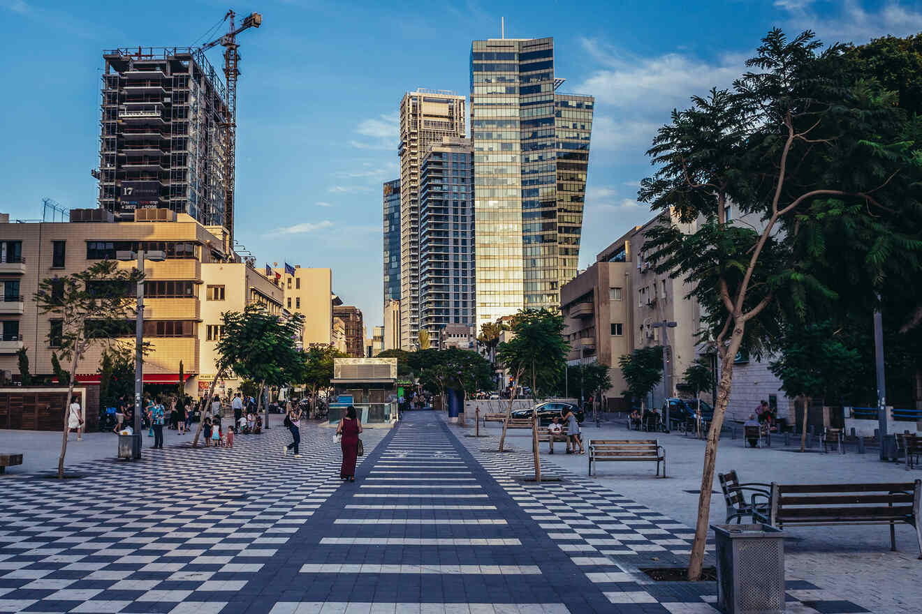 A bustling city square in Tel Aviv with pedestrians, checkerboard-patterned ground, and modern buildings under a clear sky.